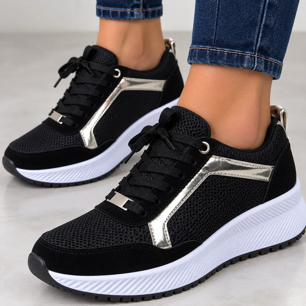 A pair of black low-top sneakers with metallic silver side panels and white platform soles, shown on a person's feet wearing blue jeans.