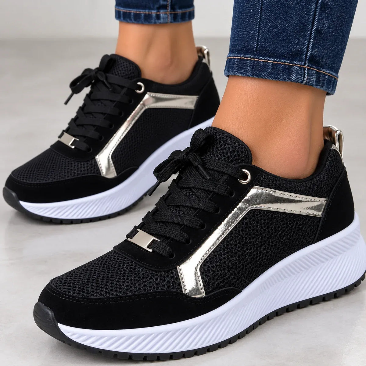 A pair of black low-top sneakers with metallic silver side panels and white platform soles, shown on a person's feet wearing blue jeans.