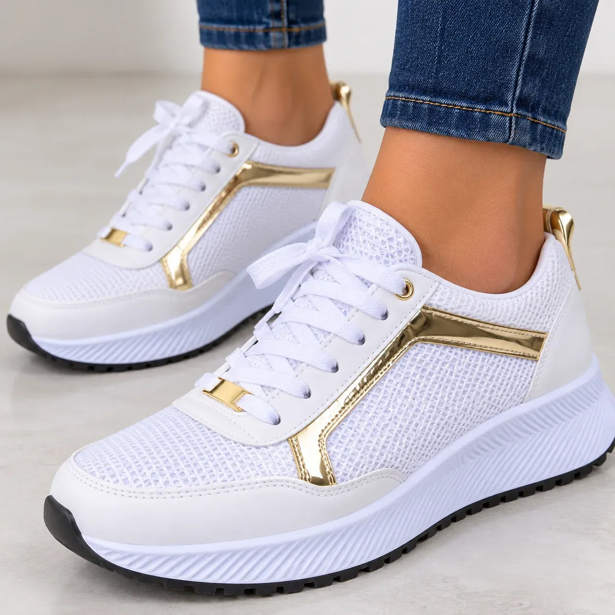 White mesh sneakers with metallic gold accents and white laces, shown on a model wearing blue jeans against a light gray background.
