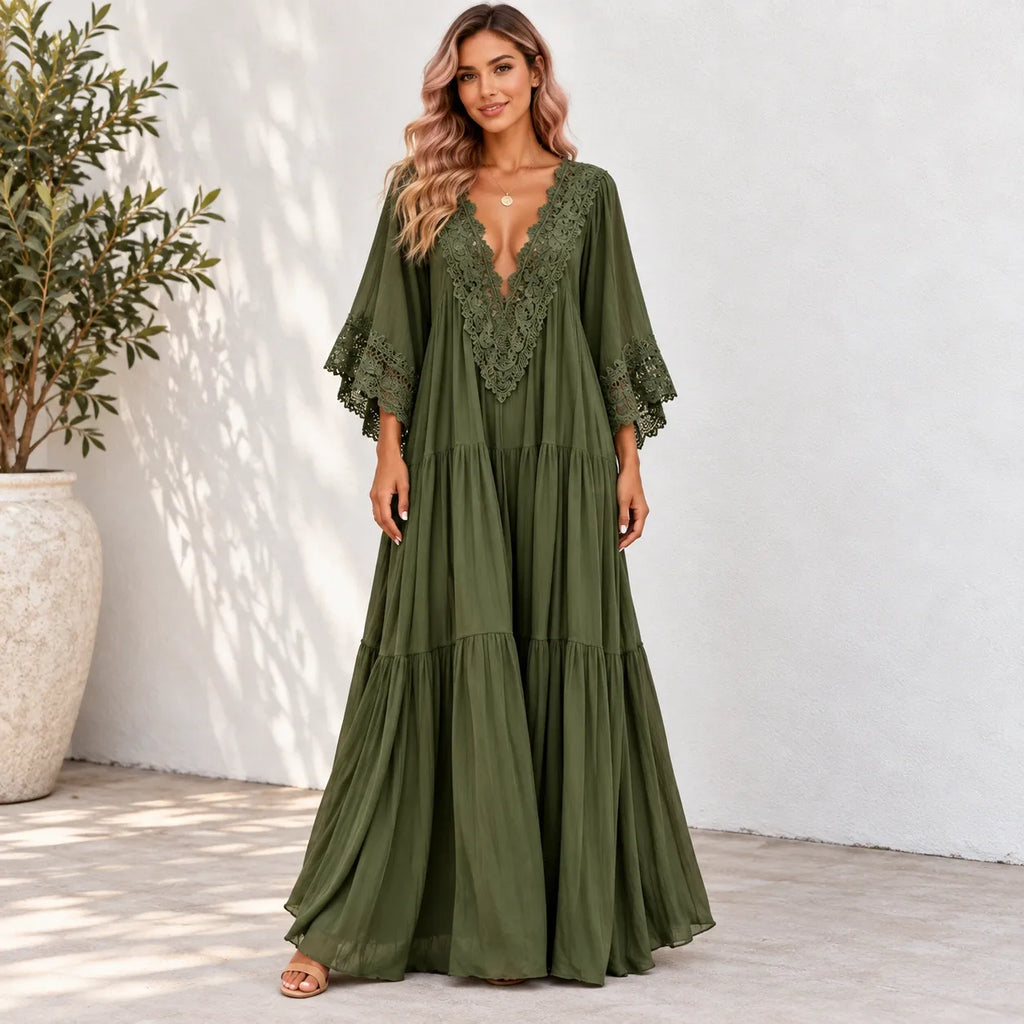 A woman models a long olive green maxi dress with a lace V-neckline and tiered skirt, standing against a white stucco wall next to a potted plant.