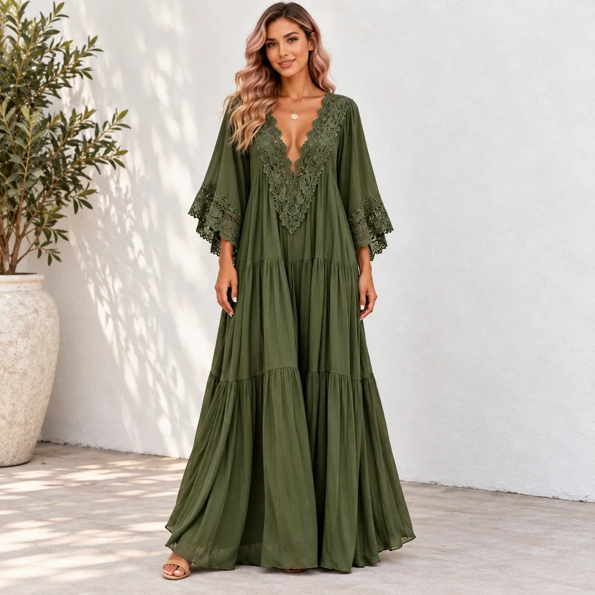 A woman models a long olive green maxi dress with a lace V-neckline and tiered skirt, standing against a white stucco wall next to a potted plant.