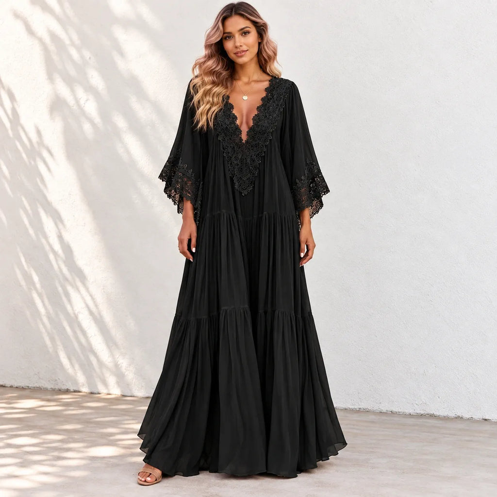 A woman modeling a black maxi dress with a deep V-neckline, lace trim, and tiered skirt against a white stucco wall with soft leaf shadows.