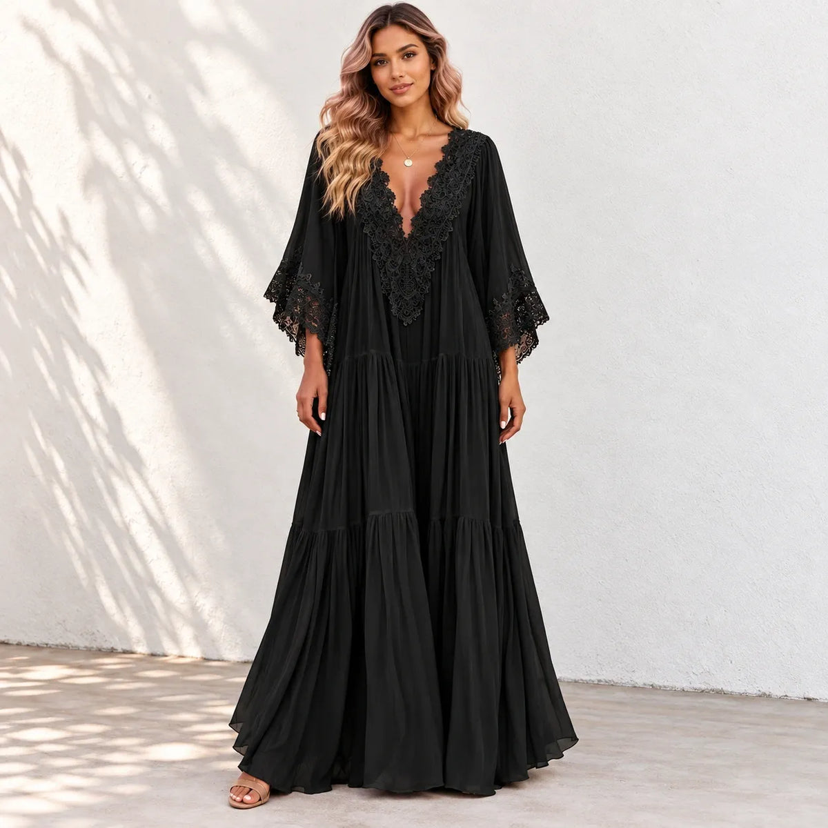 A woman modeling a black maxi dress with a deep V-neckline, lace trim, and tiered skirt against a white stucco wall with soft leaf shadows.