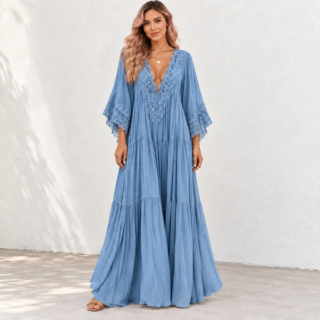 A woman modeling a light blue maxi dress with a lace V-neckline and tiered skirt, standing against a white wall.