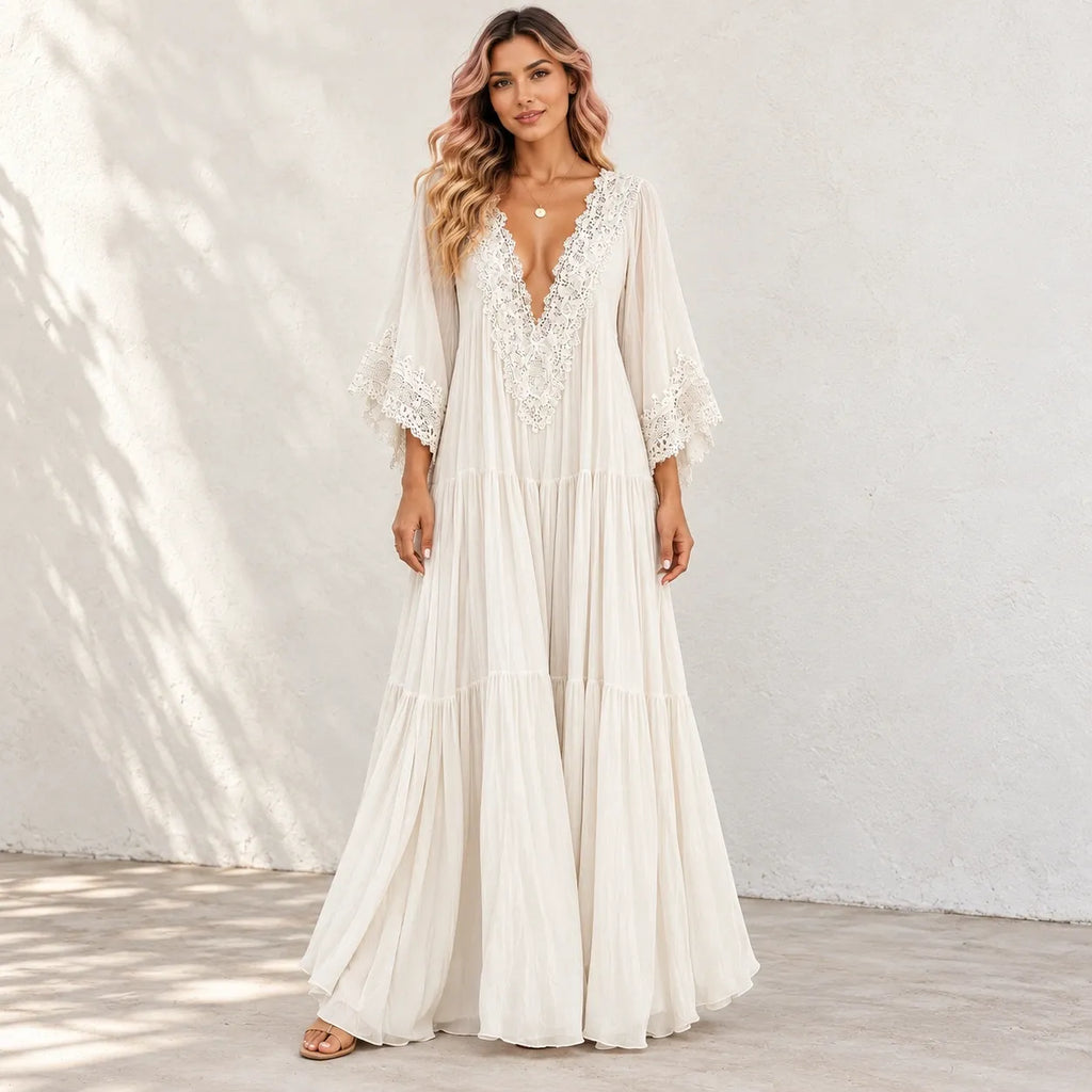 A woman models a bohemian-style maxi dress in a warm off-white/cream color. The dress features a deep V-neckline adorned with intricate lace applique, wide three-quarter length bell sleeves with lace cuffs, and a tiered, flowing skirt.