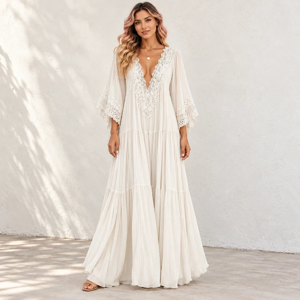 A woman models a bohemian-style maxi dress in a warm off-white/cream color. The dress features a deep V-neckline adorned with intricate lace applique, wide three-quarter length bell sleeves with lace cuffs, and a tiered, flowing skirt.