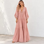 A woman modeling a dusty pink maxi dress with a lace V-neckline and lace-trimmed elbow-length sleeves, standing against a white textured wall.