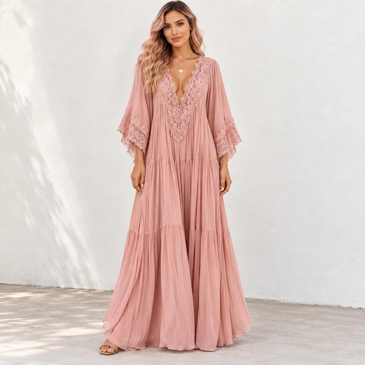 A woman modeling a dusty pink maxi dress with a lace V-neckline and lace-trimmed elbow-length sleeves, standing against a white textured wall.