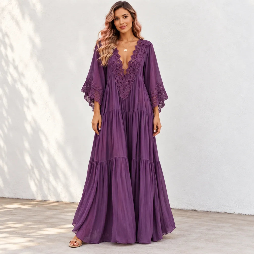 A woman wearing a long, tiered purple maxi dress with a lace V-neckline and wide bell sleeves, standing against a white stucco wall.