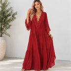 A woman models a red bohemian maxi dress with lace trim and tiered skirt in a bright, modern indoor setting with a potted plant.