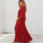 A woman modeling a long red maxi dress with flutter sleeves and lace trim, standing in a bright white room corner.