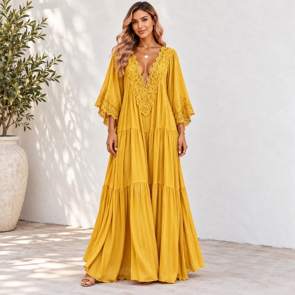 A woman models a long, flowing yellow maxi dress with a deep V-neckline, lace detailing on the bodice and sleeves, and a tiered skirt, set against a white stucco wall with a potted plant in the background.