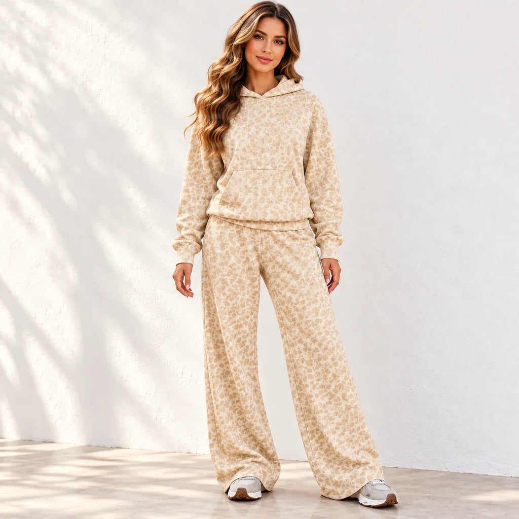 A woman modeling a matching beige and cream floral print fleece lounge set, consisting of a hooded sweatshirt and wide-leg pants, standing against a white wall with soft leaf shadows.