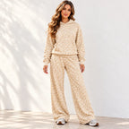 A woman modeling a matching beige and cream floral print fleece lounge set, consisting of a hooded sweatshirt and wide-leg pants, standing against a white wall with soft leaf shadows.
