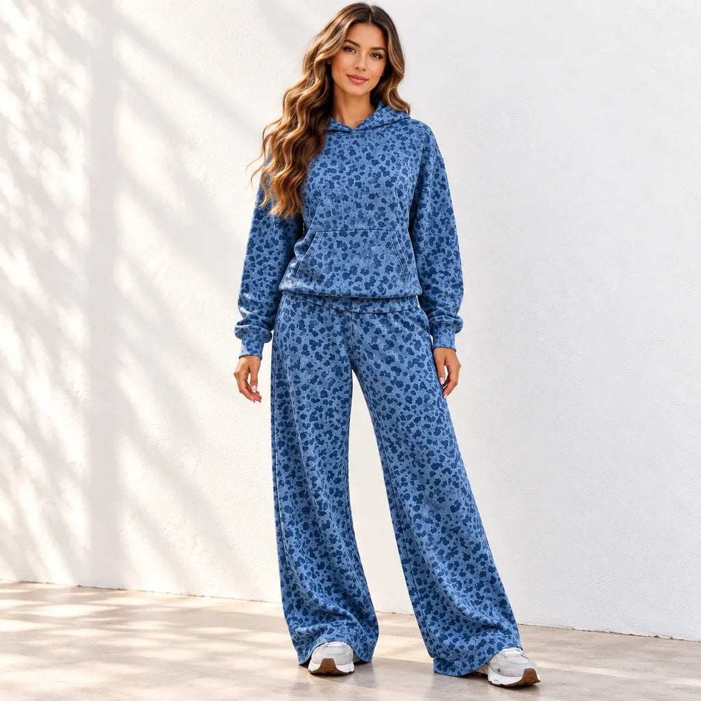 A woman stands with front to the camera wearing a matching blue leopard-print hoodie and wide-leg pants set, photographed against a white wall with soft shadow patterns.