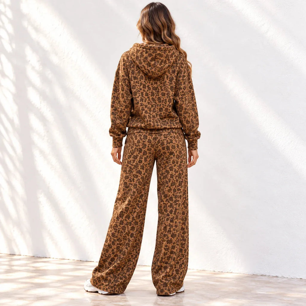 A woman stands with her back to the camera wearing a matching brown leopard-print hoodie and wide-leg pants set, photographed against a white wall with soft shadow patterns.