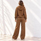 A woman stands with her back to the camera wearing a matching brown leopard-print hoodie and wide-leg pants set, photographed against a white wall with soft shadow patterns.