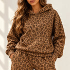 A woman wearing a brown and tan leopard-print hooded sweatshirt and matching sweatpants, photographed against a light neutral wall with soft shadows.