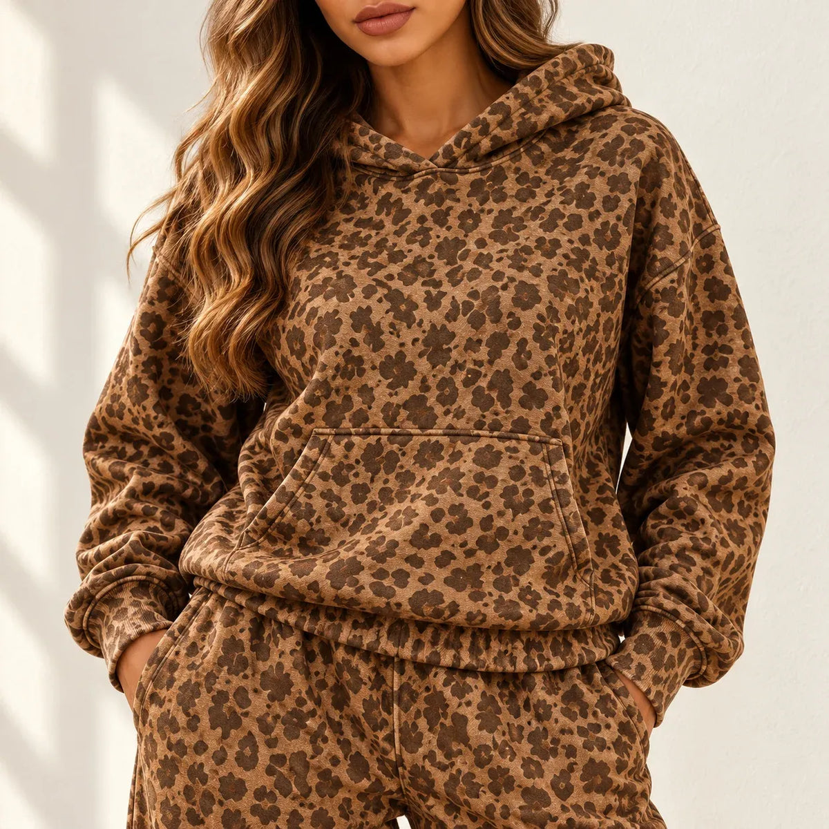 A woman wearing a brown and tan leopard-print hooded sweatshirt and matching sweatpants, photographed against a light neutral wall with soft shadows.