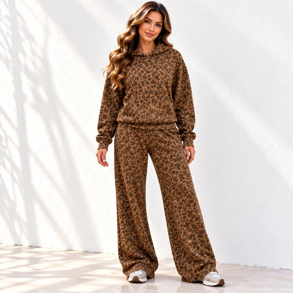 A woman stands with front to the camera wearing a matching brown leopard-print hoodie and wide-leg pants set, photographed against a white wall with soft shadow patterns.