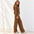 A woman stands with her side to the camera wearing a matching brown leopard-print hoodie and wide-leg pants set, photographed against a white wall with soft shadow patterns.