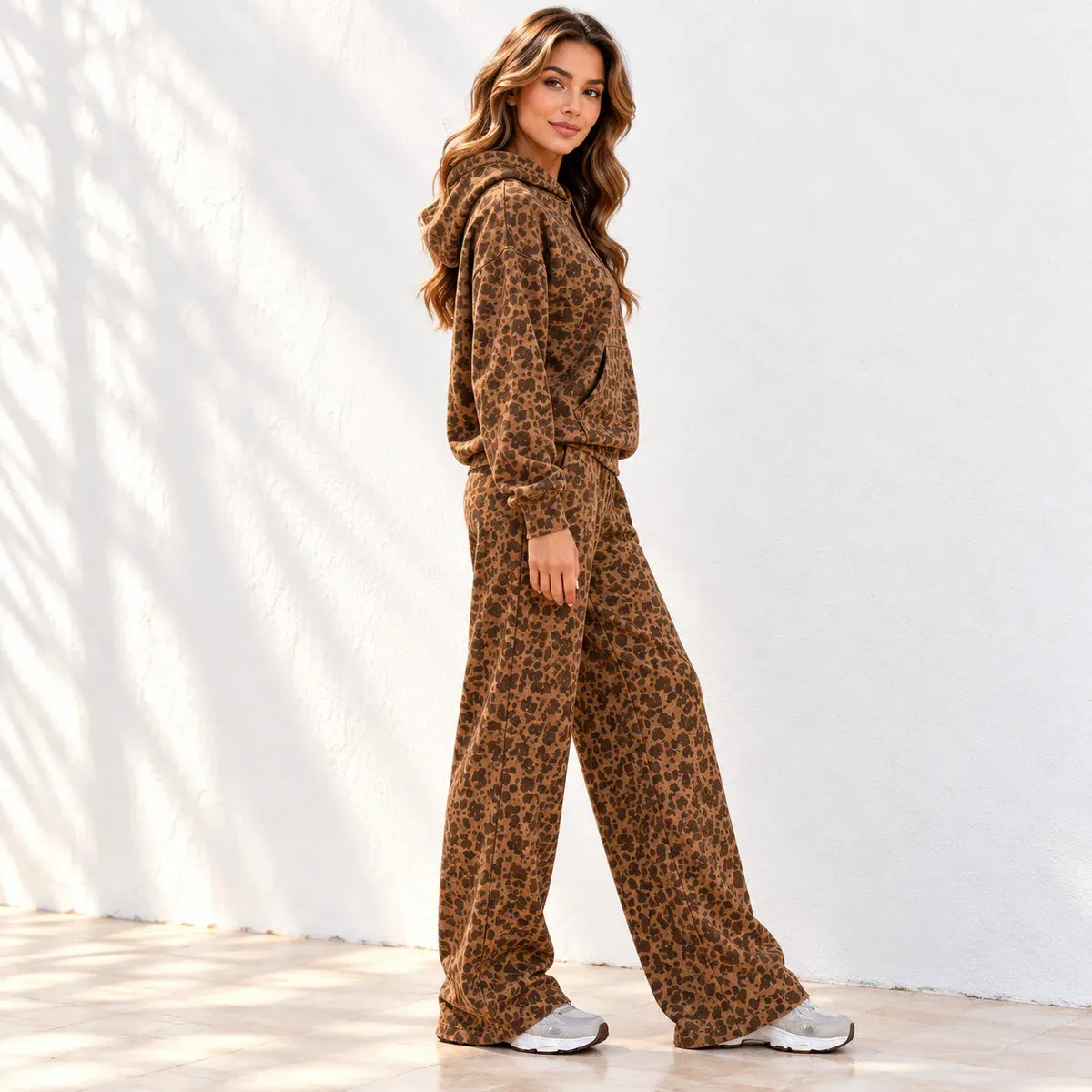 A woman stands with her side to the camera wearing a matching brown leopard-print hoodie and wide-leg pants set, photographed against a white wall with soft shadow patterns.