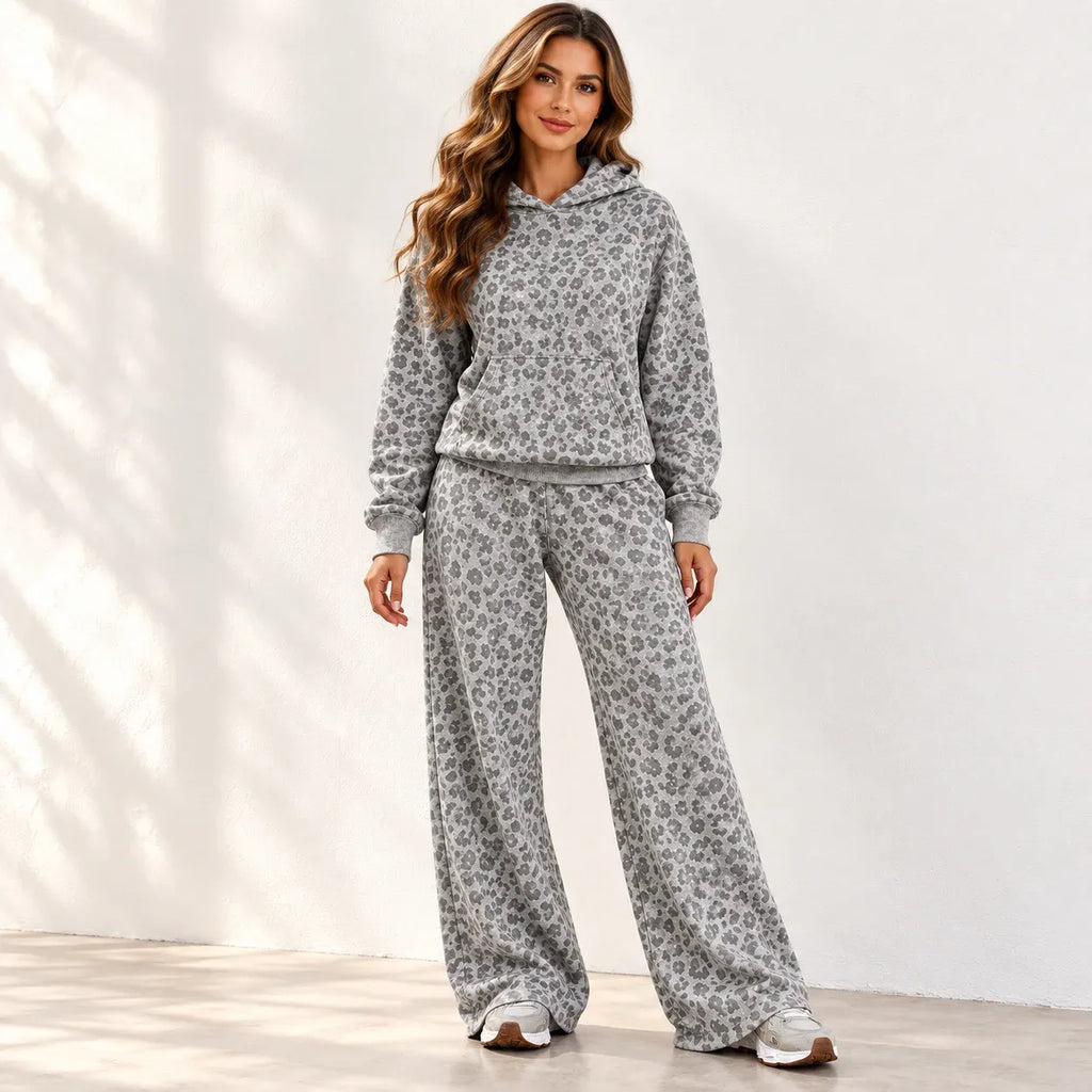 A woman modeling a matching gray and white floral-patterned loungewear set, consisting of a hooded sweatshirt and wide-leg sweatpants, standing against a white wall with soft shadow patterns.