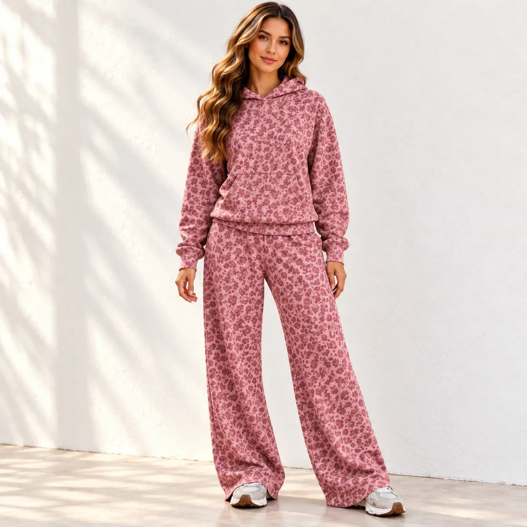 A woman stands against a white wall wearing a matching pink floral hoodie and wide-leg sweatpants set, with white sneakers. The background has soft leaf shadows cast across it.