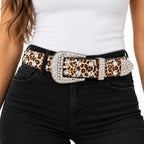 A close-up of a model wearing a leopard print belt with a large silver-tone buckle embellished with rhinestones, styled with a white ribbed top and black jeans.