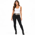A woman modeling black skinny jeans with a decorative metallic patterned belt, a white tank top, and black ankle boots, photographed against a solid white background.