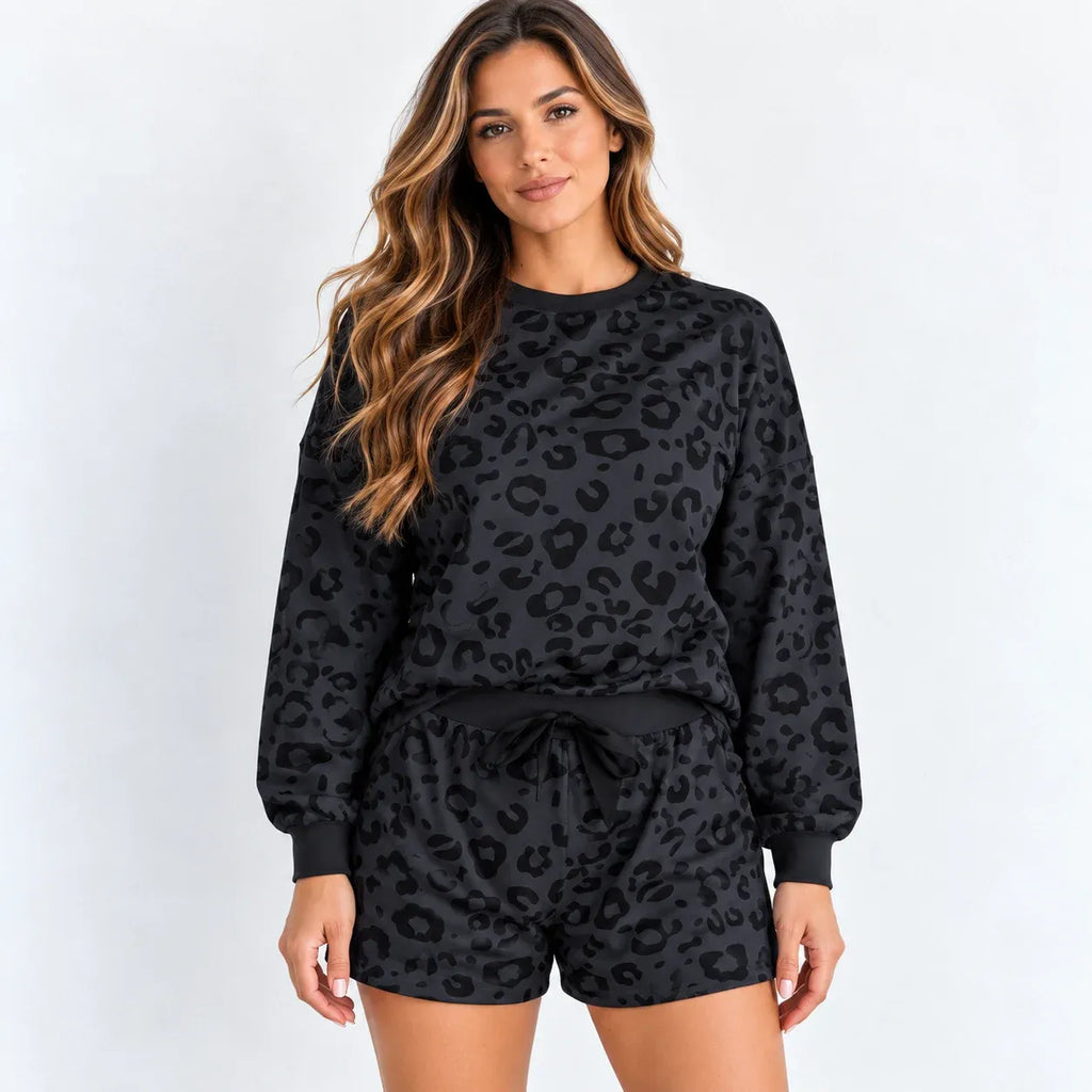A woman modeling a matching black leopard-print lounge set, featuring a long-sleeve sweatshirt and drawstring shorts, against a plain white studio background.