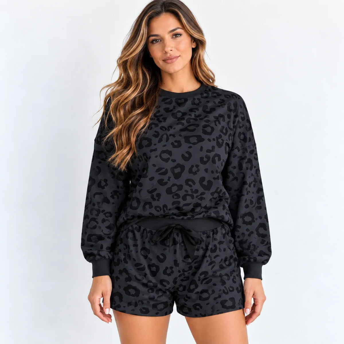 A woman modeling a matching black leopard-print lounge set, featuring a long-sleeve sweatshirt and drawstring shorts, against a plain white studio background.
