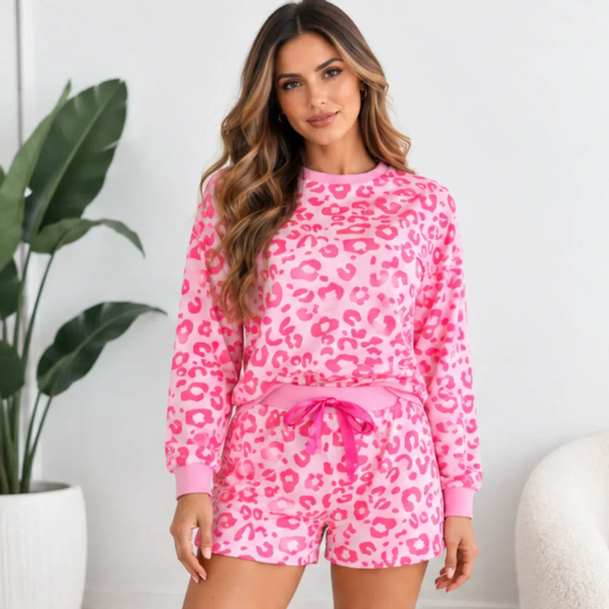 A woman modeling a pink long-sleeve sweatshirt and matching shorts set with a darker pink leopard print pattern, standing in a bright indoor setting with a potted plant and white chair visible.