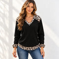 A woman wearing a black long-sleeve waffle-knit top with leopard print trim on the V-neck, cuffs, and hem, paired with blue jeans, standing against a white wall with soft leaf shadows.