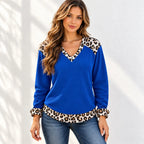 A woman wearing a royal blue long-sleeve knit top with leopard print trim at the V-neck, cuffs, and hem, paired with blue jeans, standing against a white wall with soft leaf shadows.