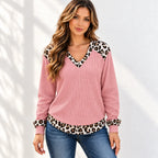 A woman wearing a pink waffle-knit long-sleeve sweater with leopard print trim on the V-neck, cuffs, and hem, paired with blue jeans against a white wall with soft leaf shadows.