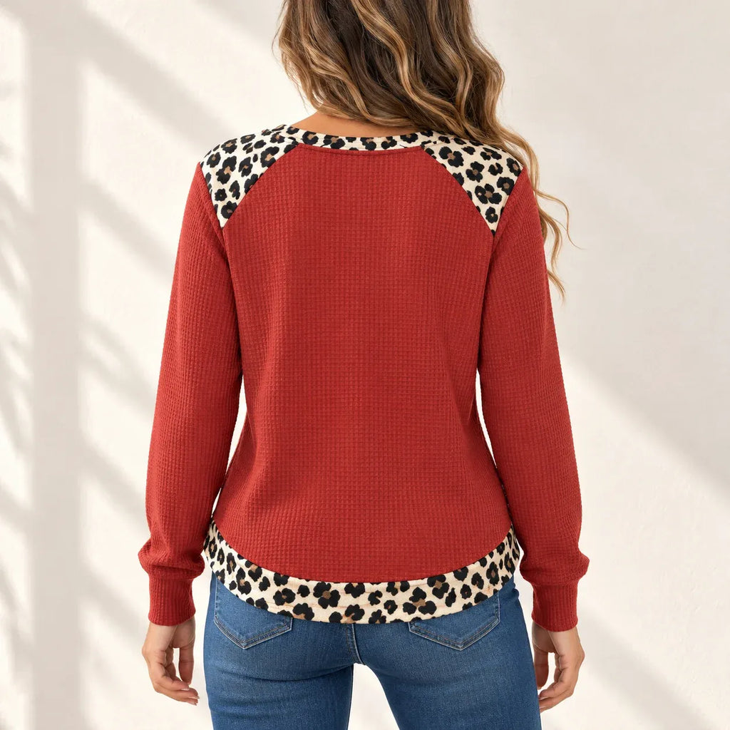 A woman shown from the back wearing a rust red waffle-knit sweater with leopard print trim along the shoulders and hem, paired with blue jeans against a white wall with soft shadow patterns.
