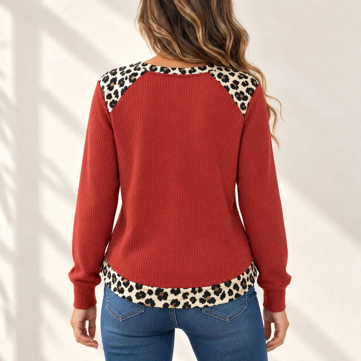 A woman shown from the back wearing a rust red waffle-knit sweater with leopard print trim along the shoulders and hem, paired with blue jeans against a white wall with soft shadow patterns.