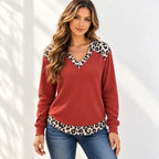 A woman wearing a rust red waffle-knit sweater with leopard print trim on the shoulders and hem, paired with blue jeans, standing against a white wall with soft leaf shadows.
