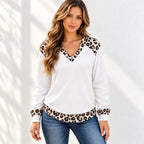 A woman wearing a white waffle-knit long-sleeve top with leopard print trim on the V-neck, cuffs, and hem, paired with blue jeans, standing against a white wall with soft leaf shadows.