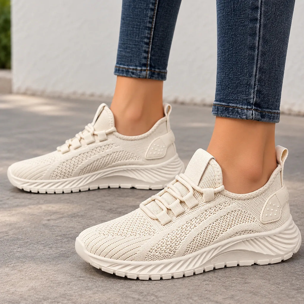 A pair of women's knit slip-on sneakers in a light cream/off-white color, shown being worn with blue jeans on a gray concrete floor.