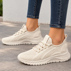 A pair of women's knit slip-on sneakers in a light cream/off-white color, shown being worn with blue jeans on a gray concrete floor.