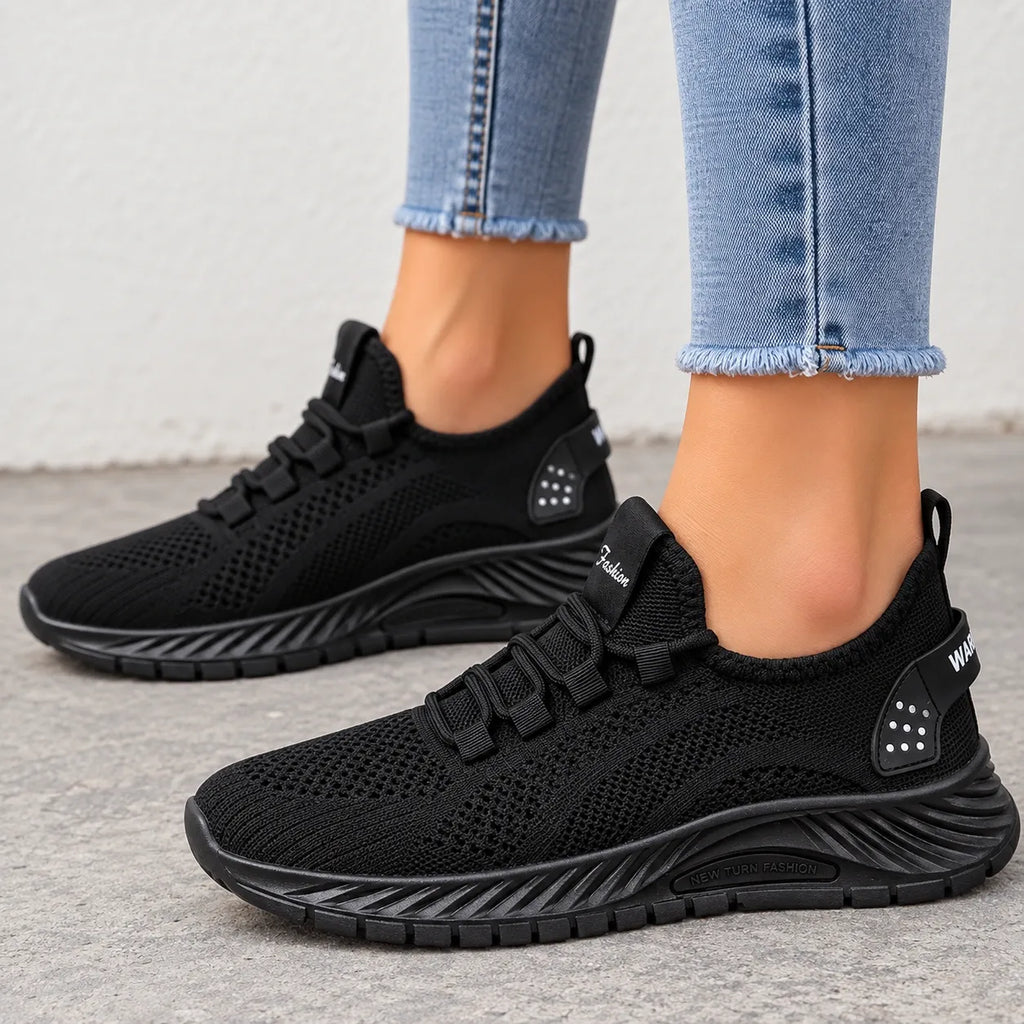 A pair of black slip-on mesh sneakers worn with light blue frayed-hem jeans, shown from the side on a gray concrete floor.