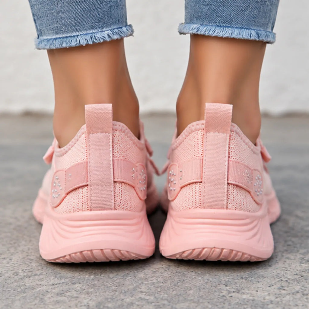 A close-up rear view of a person wearing light pink mesh sneakers with hook-and-loop strap details and small rhinestone embellishments, paired with frayed blue jeans.