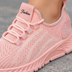 A close-up of a light pink knit slip-on sneaker being worn, with a soft fabric tongue patch reading 'Fashion' in black script. The shoe has matching pink laces, a textured knit upper, and a chunky molded sole, shown against a gray stone-like surface.
