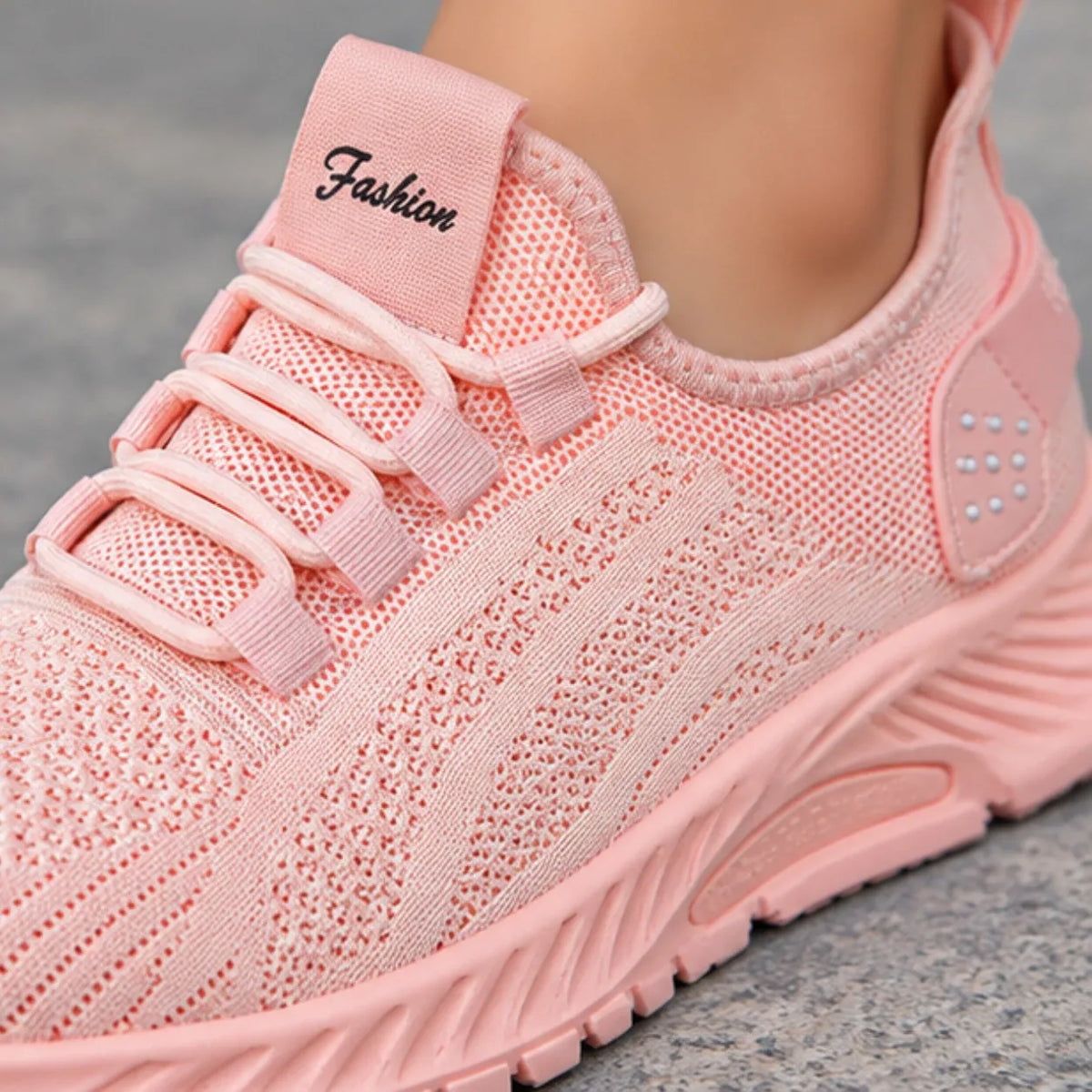 A close-up of a light pink knit slip-on sneaker being worn, with a soft fabric tongue patch reading 'Fashion' in black script. The shoe has matching pink laces, a textured knit upper, and a chunky molded sole, shown against a gray stone-like surface.