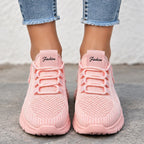 A pair of light pink knit sneakers with matching pink laces, shown on a person wearing light blue frayed-hem jeans. The shoes feature a textured knit upper, a pink tongue patch with the word 'Fashion' printed on it, and thick cushioned soles, photographed against a neutral concrete and stucco background.