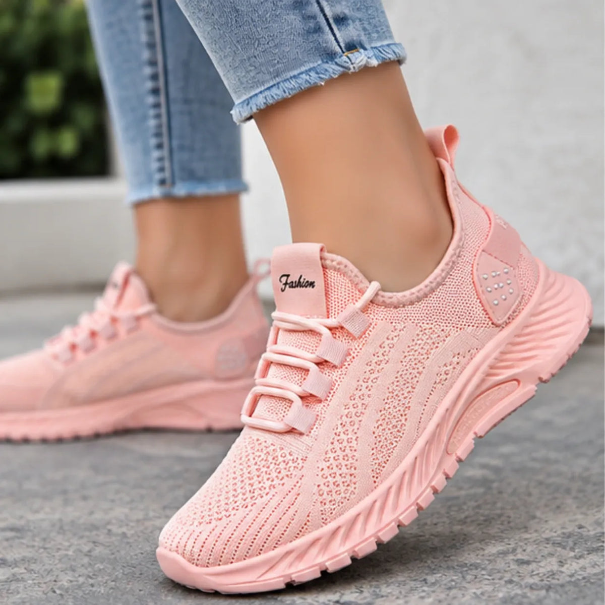 A person wearing light pink knit athletic sneakers with matching laces and a thick cushioned sole, paired with light blue jeans, standing on a gray concrete surface.
