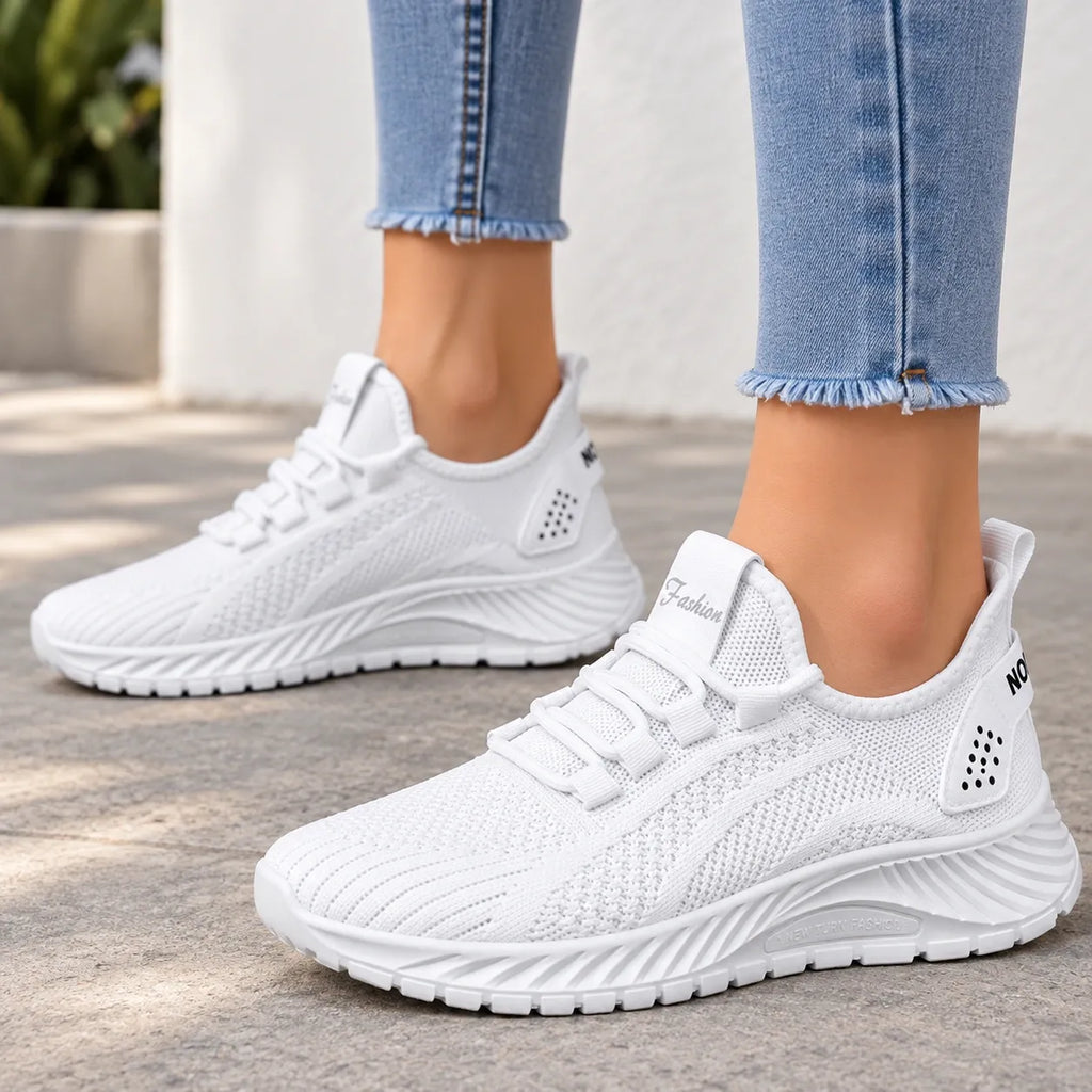 A pair of white knit slip-on sneakers worn with light blue frayed-hem jeans, shown on a person's feet against a light concrete and wall background.