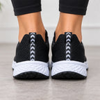 Black knit athletic sneakers with white chunky soles, shown from the back on a person's feet against a light gray indoor floor.